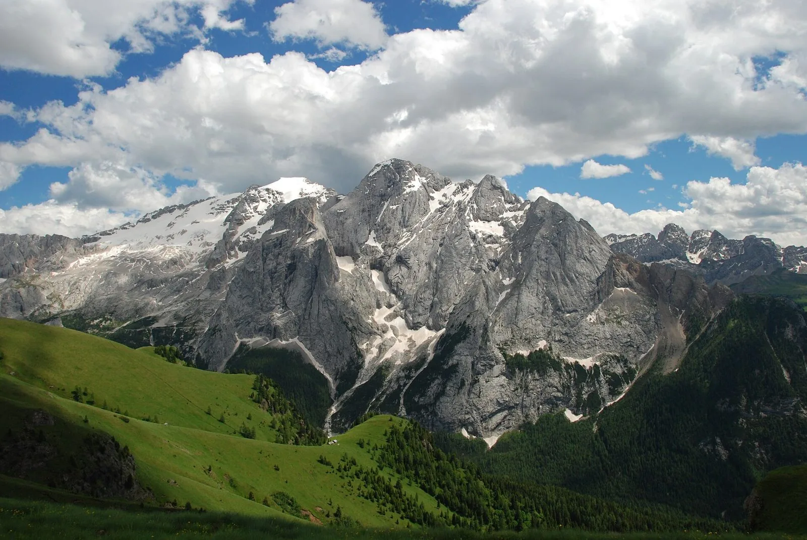 Il massiccio della Marmolada con il ghiacciaio innevato a sinistra e grandi pareti di roccia grigia, pascoli verdi e boschi in primo piano, cielo azzurro con nuvole bianche