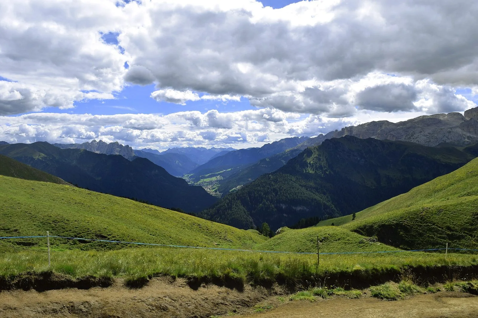 Vista dall'alto su una valle verde delle Dolomiti con un paese sul fondovalle, pascoli in primo piano, boschi scuri e pareti rocciose sulla destra sotto un cielo nuvoloso