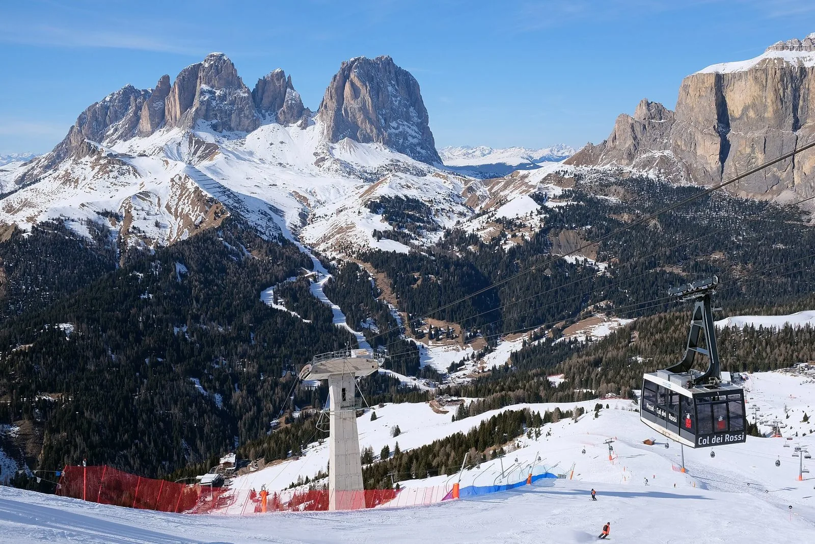 Pista da sci innevata con alcuni sciatori lontani, una cabina della funivia Col dei Rossi e un pilone; sullo sfondo le guglie del Sassolungo e le pareti del Gruppo del Sella sotto un cielo azzurro