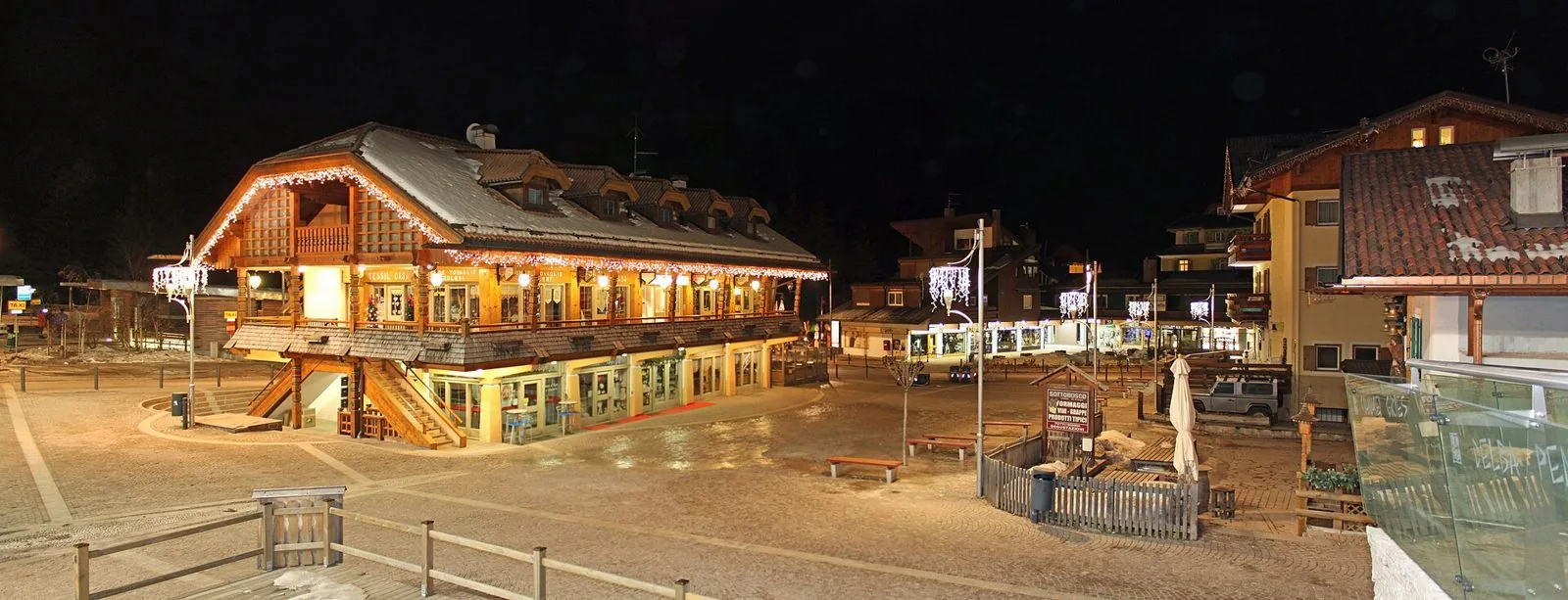 Piazza di Canazei di notte con un grande edificio in legno decorato da luminarie natalizie, lampioni con luci bianche e selciato leggermente innevato