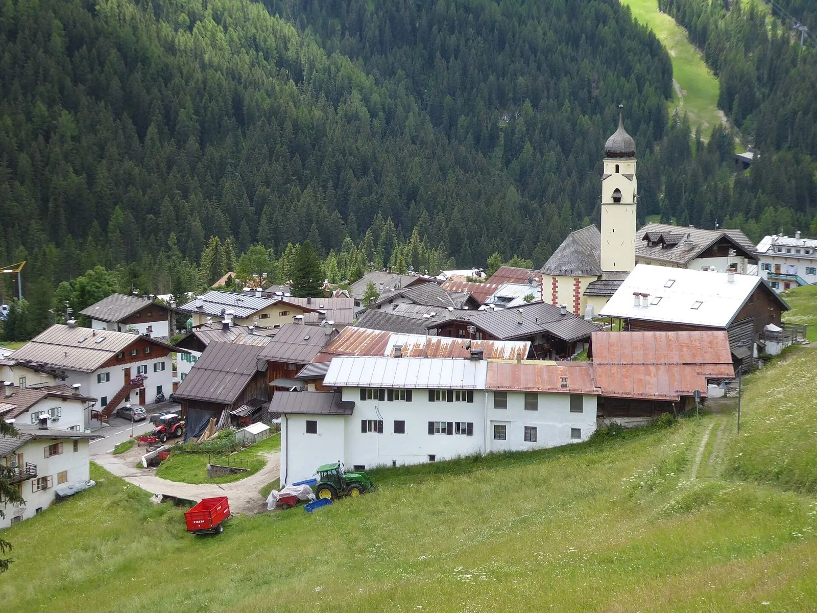 Il piccolo borgo di Penia con case bianche, tetti in lamiera e il campanile a cipolla, circondato da prati e da un ripido bosco di abeti