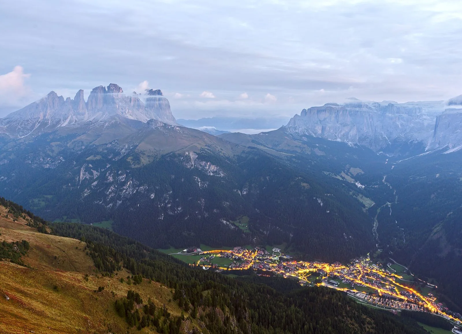Veduta dall'alto di un paese illuminato sul fondovalle alle prime luci del giorno, tra boschi di conifere, con le guglie del Sassolungo a sinistra e le pareti del gruppo del Sella a destra avvolte da nuvole basse