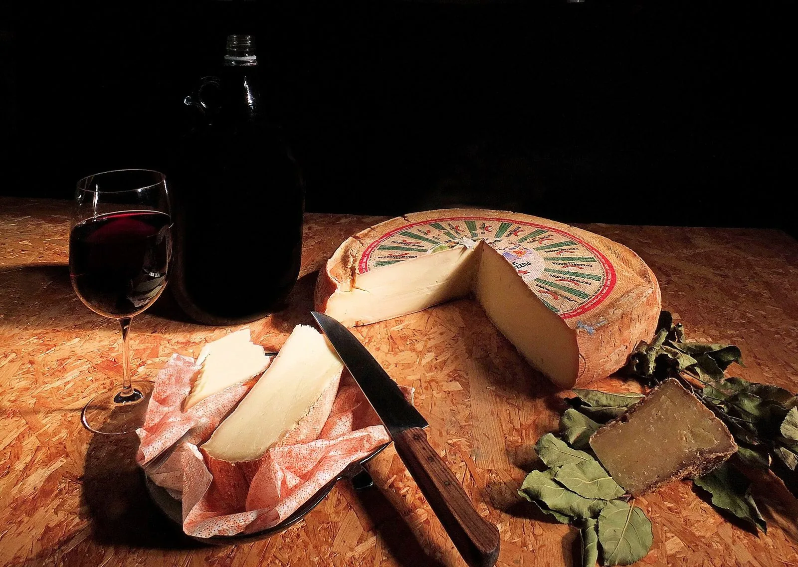 Forma di Puzzone di Moena con uno spicchio tagliato, fette di formaggio su carta, un coltello, un calice di vino rosso, una bottiglia scura e foglie di alloro su un tavolo di legno