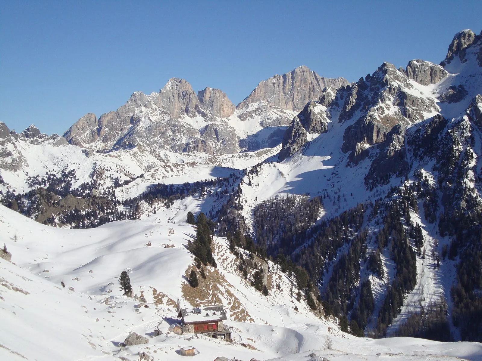 Un rifugio in legno e pietra con le imposte rosse in mezzo a pendii coperti di neve, boschi di conifere e cime dolomitiche rocciose sotto un cielo blu terso