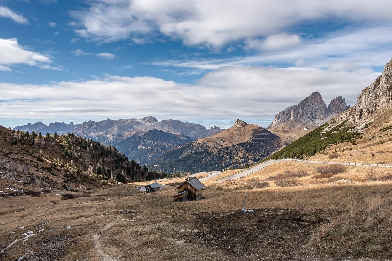 Strada di montagna che attraversa pascoli d'alta quota dai colori autunnali, con due baite in legno e le cime del Sassolungo sullo sfondo sotto un cielo nuvoloso